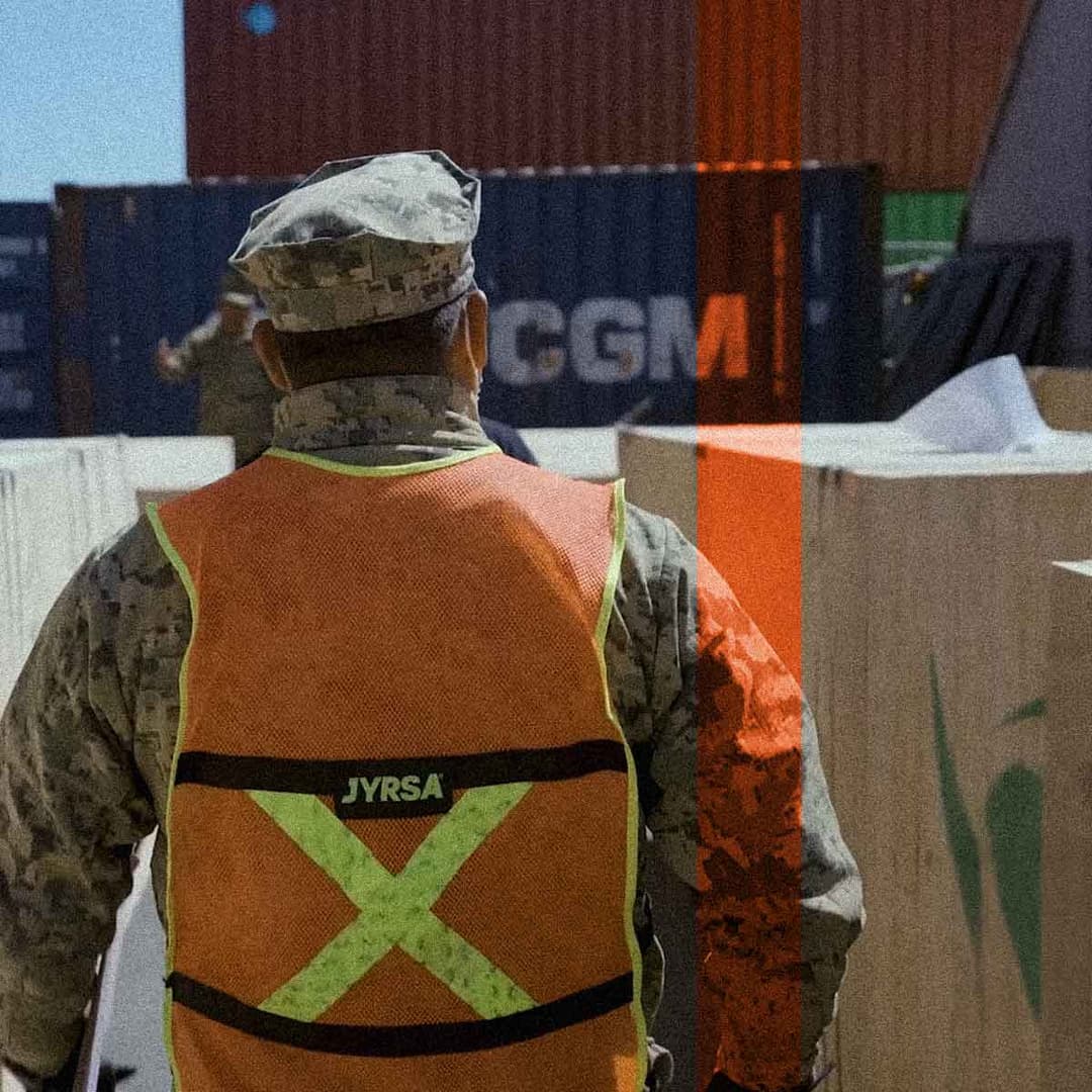 Una persona vestida con uniforme militar y un chaleco de seguridad naranja camina entre grandes cajas y contenedores de transporte en una zona industrial al aire libre, inspeccionando en busca de posibles casos de fraude.