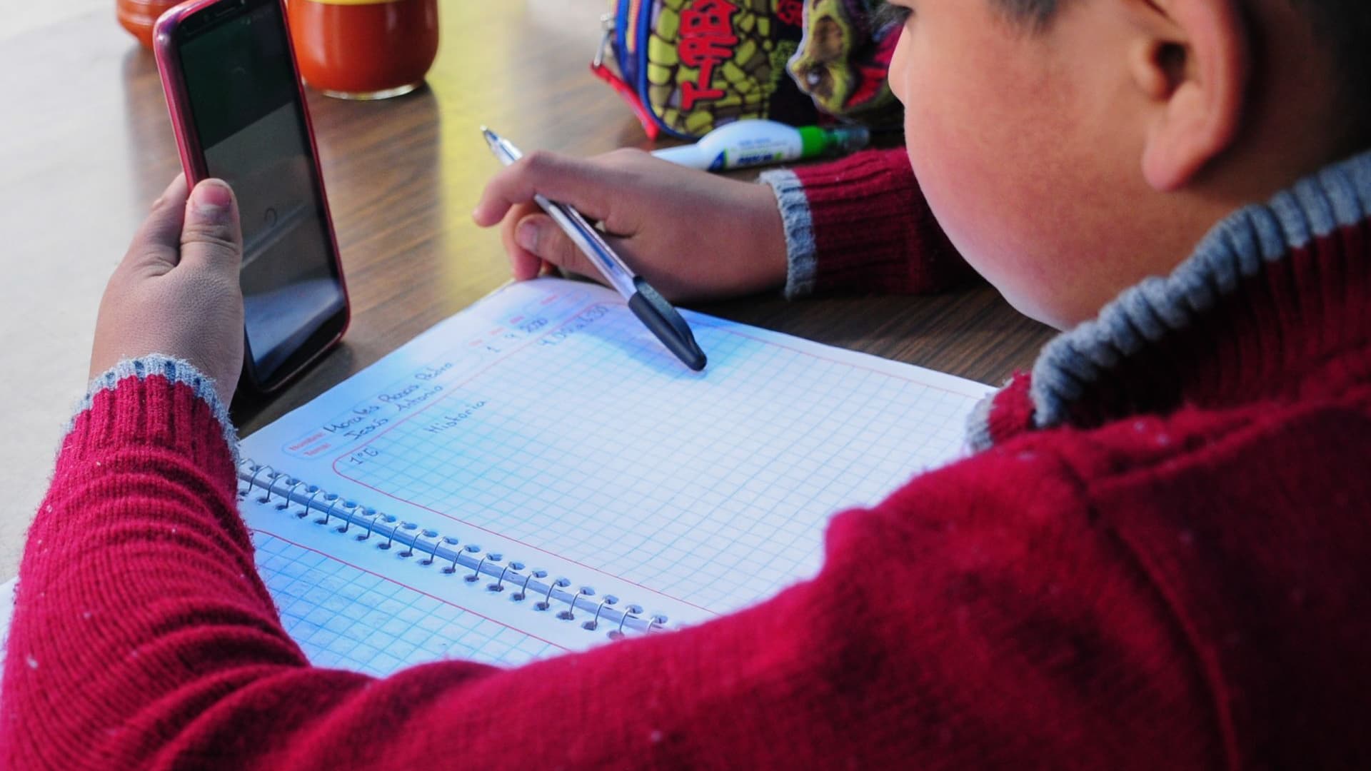 Un niño escribe en un cuaderno y ve un celular