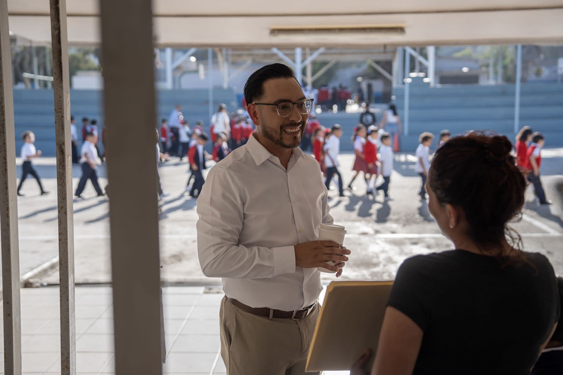 Dos personas hablan en una escuela con folders en la mano y niños de fondo