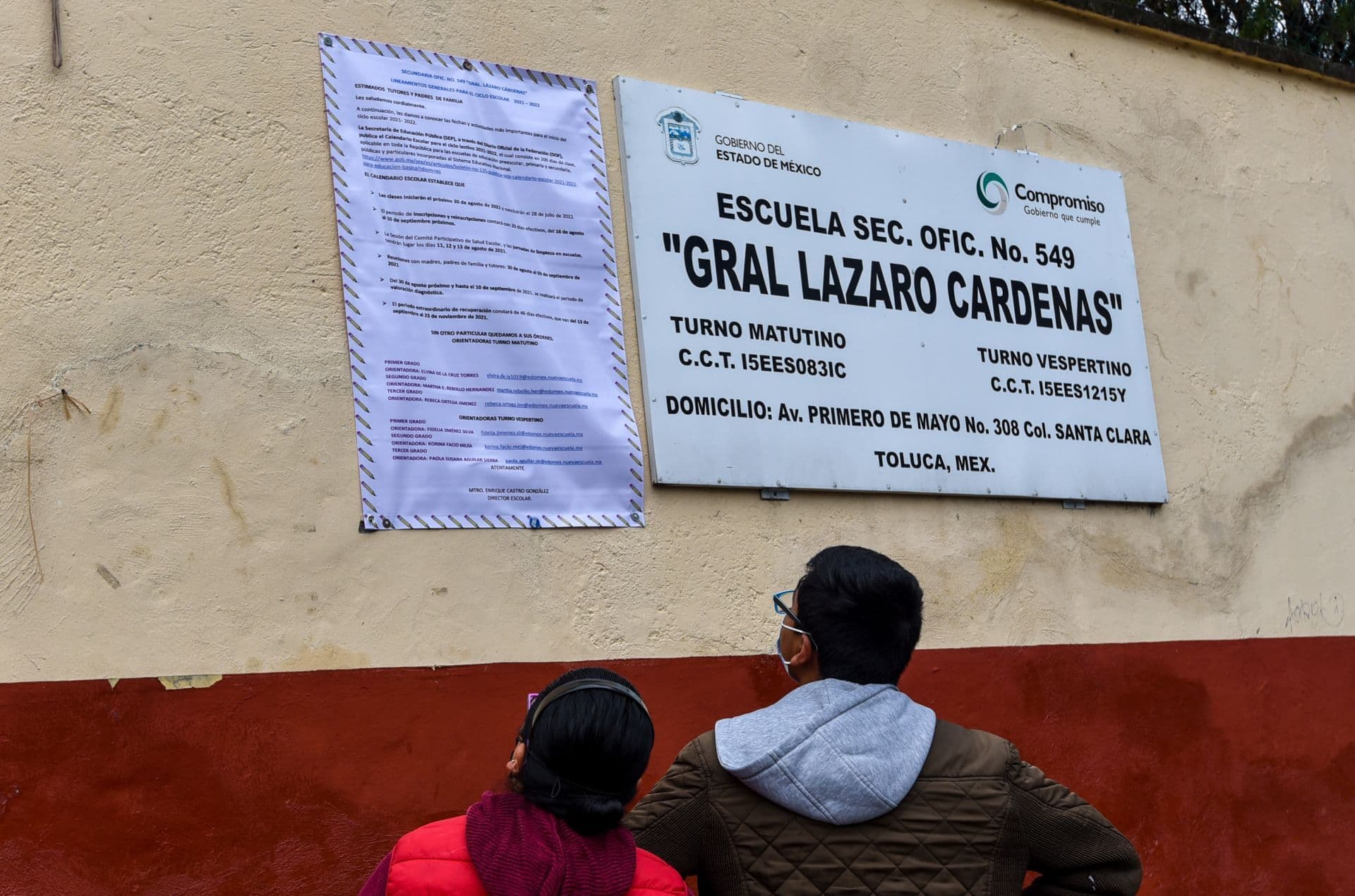 Dos personas revisan indicaciones afuera de una escuela, que incluye la Carta de Asignación de la SEP