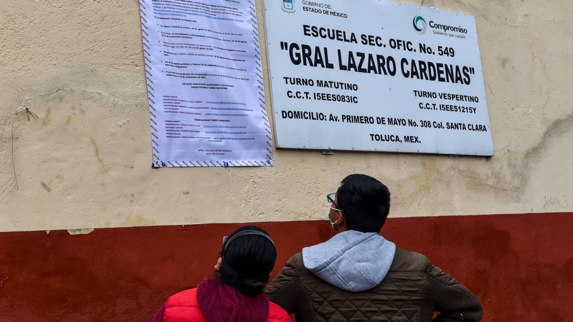 Dos personas revisan indicaciones afuera de una escuela, que incluye la Carta de Asignación de la SEP