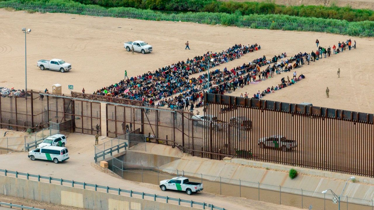 Grupo de migrantes esperando en la frontera entre México y Estados Unidos, con vehículos y vallas de seguridad presentes.