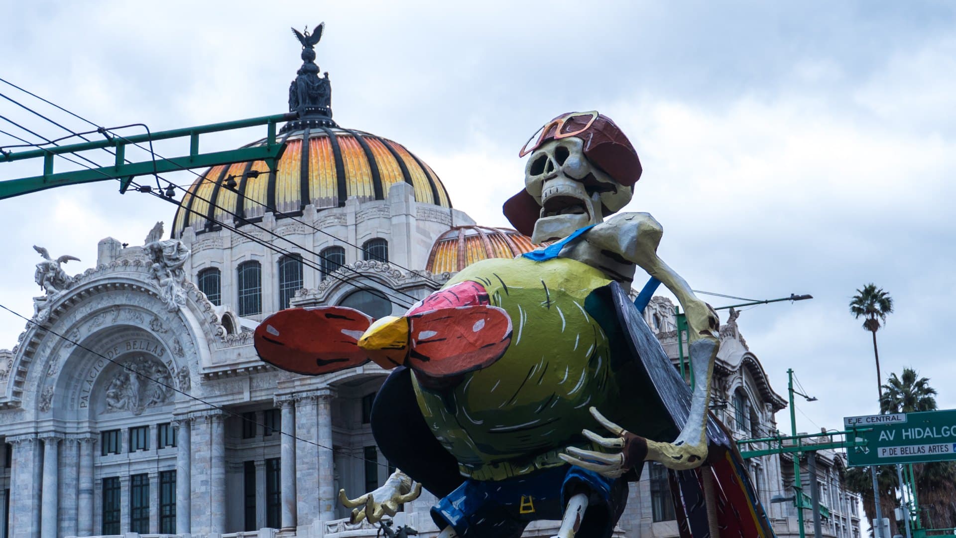 Una calavera con gorra y lentes sostiene un ave de colores frente a la cúpula del Palacio de Bellas Artes. Foto: N+